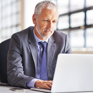 Smaller image of middle-aged Caucasian man in business attire sits at desk working on laptop