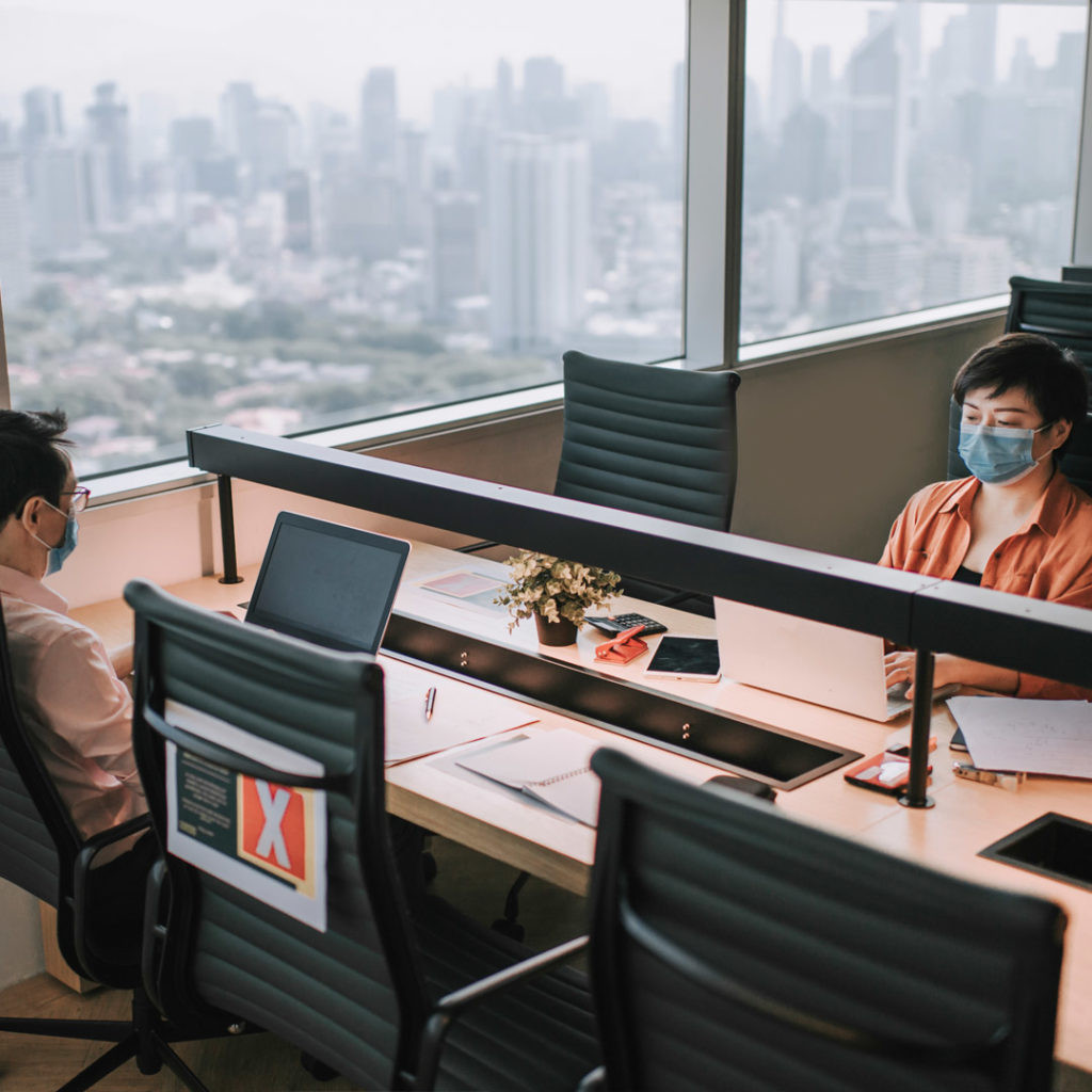 Image of two office workers sitting adjacent from each other wearing masks working on laptops with city in the background
